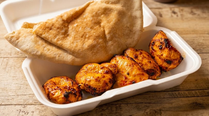 Grilled chicken pieces and pita bread in a white takeout container on a wooden table.