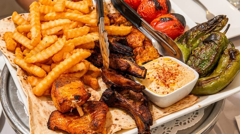 A platter of grilled meats, french fries, roasted vegetables, and hummus on flatbread.