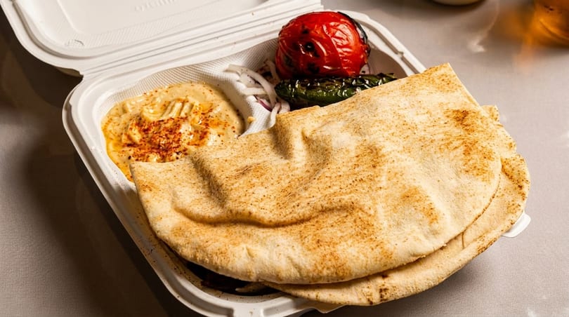 Takeout container with hummus, pita bread, grilled tomato, green pepper, and sliced onions.